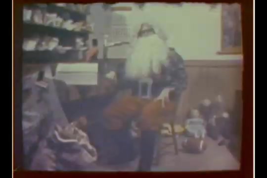 The still depicts Santa Claus seated in a cozy department store setting, with a traditional backdrop. He wears a classic red suit and a long white beard. Nearby, shelves display various items, suggesting a festive atmosphere. Toys and other decorations are visible, hinting at the store's holiday theme. The image captures the essence of the 1970s holiday spirit as part of a CBS News story on innovative Santa visits.
