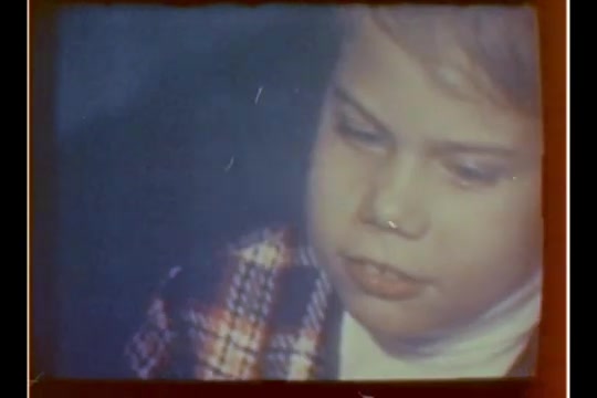 The 16mm still features a young child with a focused expression, possibly in a moment of contemplation or anticipation. The child is wearing a plaid jacket over a white shirt, and the overall color tone indicates a warm, nostalgic atmosphere typical of holiday-themed imagery. The blurred background suggests a festive or busy environment, likely related to a department store visit to Santa.
