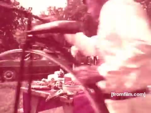 The still from the 16mm Pabst Beer commercials features a bustling outdoor scene. A person is focused on a grill, presumably preparing food, while a picnic table loaded with various dishes can be seen nearby. The atmosphere suggests a casual, social gathering with a vintage feel typical of the 1970s. The colors have a faded quality, characteristic of older film prints.