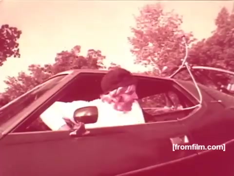 The 16mm still from the 1970s Pabst Beer commercials depicts an individual in a car. The scene features a vintage vehicle with distinctive styling of the era. The person is likely engaging in a casual or humorous moment, fitting the light-hearted tone typical of commercials from that time. The overall color palette may have a warm, slightly faded look, characteristic of older film stock. This captures the nostalgic vibe of 1970s American advertising.