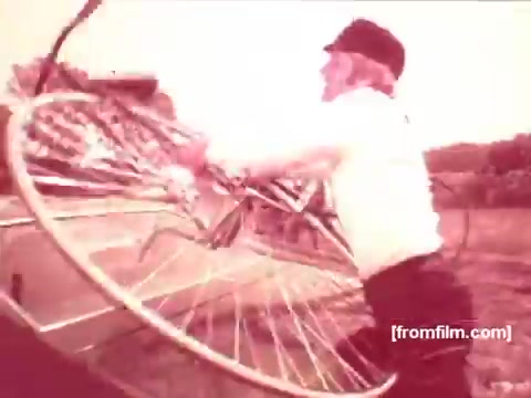 The still depicts a scene from a 1970s Pabst Beer commercial, featuring a man in a cap and white shirt handling a bicycle. The color tone is warm and faded, characteristic of 16mm film, suggesting a nostalgic or vintage vibe. The setting appears outdoors, likely reflecting a carefree, casual atmosphere associated with beer enjoyment.
