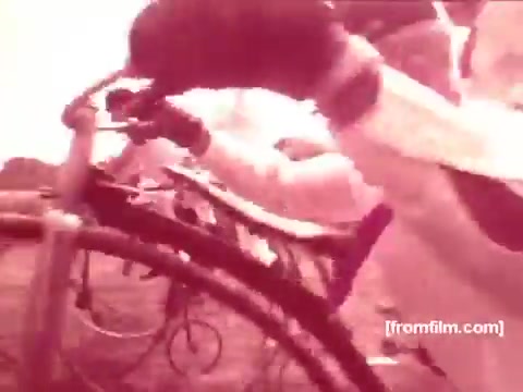 The still from the 1970s Pabst Beer commercials features a close-up of a bicycle, focusing on someone's hands as they grip the handlebars. The image has a vintage feel, typical of 16mm film, with a reddish tint that suggests a nostalgic ambiance. The setting appears outdoor, hinting at an active, perhaps playful lifestyle associated with the brand. The overall aesthetic emphasizes a carefree, spirited vibe often marketed in beer commercials of that era.