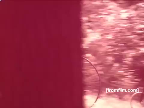 The still from the 'Pabst Beer Commercials - 1970's' shows a muted reddish tint, likely a result of the film stock or lighting conditions. In the scene, a natural setting is partially visible with blurred greenery in the background, suggesting a casual, outdoor atmosphere typical of beer commercials from that era. The visual elements evoke a nostalgic feel, recalling the carefree vibes of 1970s advertising.