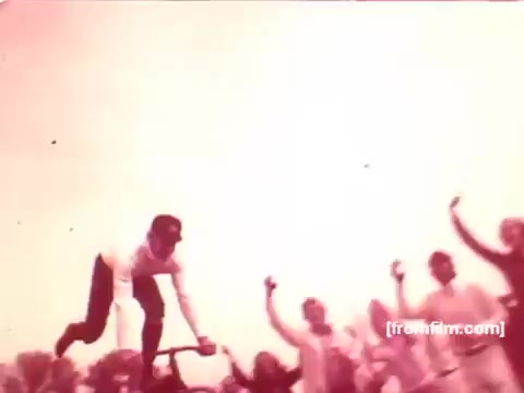 The still features a vibrant scene from a 1970s Pabst Beer commercial. A man, possibly enjoying a celebratory moment, is depicted mid-action on a bicycle, appearing to interact with a cheering crowd. The overall atmosphere is lively and festive, emphasizing camaraderie and celebration associated with the beer brand. The visual quality reflects the characteristics of 16mm film, capturing the nostalgic essence of that era.