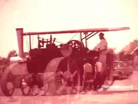 The still from the 'Pabst Beer Commercials - 1970s' features a vintage scene involving an agricultural setting, likely showcasing machinery like a steam engine or tractor. In the background, a few individuals are engaged around the machinery, while classic cars can be seen parked, hinting at the era's aesthetics. The overall tone is nostalgic, reflecting the rustic charm often associated with commercials from that time promoting Pabst Beer.