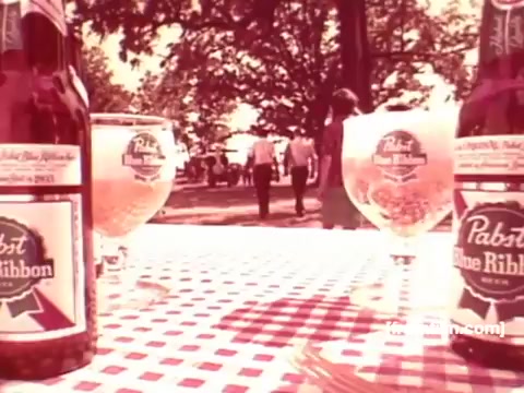 The still features a picnic setting with two bottles and glasses of Pabst Blue Ribbon beer prominently displayed on a checkered tablecloth. In the background, a couple of people stroll through a park-like area under the shade of trees. The scene evokes a casual, social atmosphere typical of 1970s beer commercials, emphasizing enjoyment and camaraderie. The overall tone is nostalgic and light-hearted.