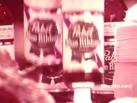 The still from the 16mm 'Pabst Beer Commercials - 1970's' features bottles of Pabst beer prominently displayed, likely in a social or bar setting. The scene captures the vintage aesthetic typical of that era, with a warm color palette and an emphasis on the branding of the beer. It suggests a casual, celebratory atmosphere, reflecting the marketing style of the time that aimed to associate the product with enjoyable social moments.