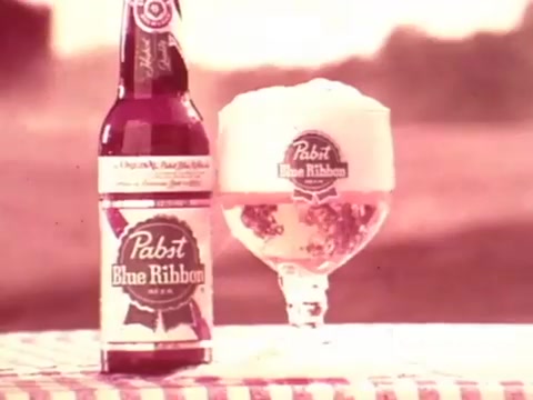 The still features a bottle and a glass of Pabst Blue Ribbon beer. The bottle stands upright next to a full glass, which has a frothy head. The background suggests an outdoor setting, and the scene has a soft, vintage aesthetic typical of 1970s commercials. The tablecloth is checkered, adding to the nostalgic vibe of the era. Overall, it conveys a relaxed, social drinking experience associated with the brand.