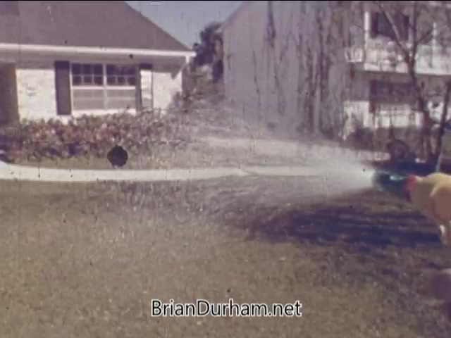The still from the 'Partial Ronco Spray Gun Commercial - 1960s' shows a person using a colorful spray gun in a residential yard. Water is forcefully spraying out, creating a wide mist effect. The background features a house and landscaped garden, typical of suburban settings in that era. The scene captures a lively demonstration of the product, likely aimed at showcasing its effectiveness for home or garden use.