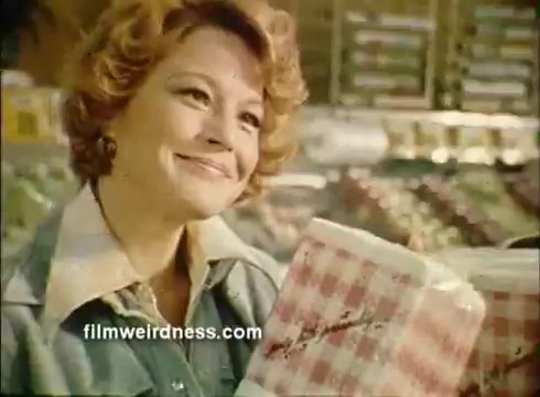The still features a smiling woman with curly hair, holding a pack of Mardi Gras paper napkins in a grocery store. She is dressed in a light-colored jacket, showcasing a cheerful demeanor. The background displays shelves filled with various products, evoking a lively shopping atmosphere typical of commercials from that era. The overall tone is inviting and festive, aligned with the Mardi Gras theme.