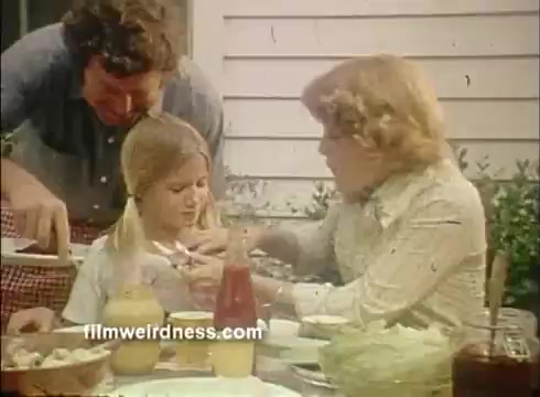 The still depicts a family meal scene featuring a young girl at the center, sitting at a table filled with food. A man, possibly her father, serves her from a dish, while a woman, likely her mother, assists her with a condiment. The setting appears casual, with a homey atmosphere, showcasing the interaction and care typical of family gatherings, highlighting the playful and festive spirit associated with the use of Mardi Gras paper napkins.