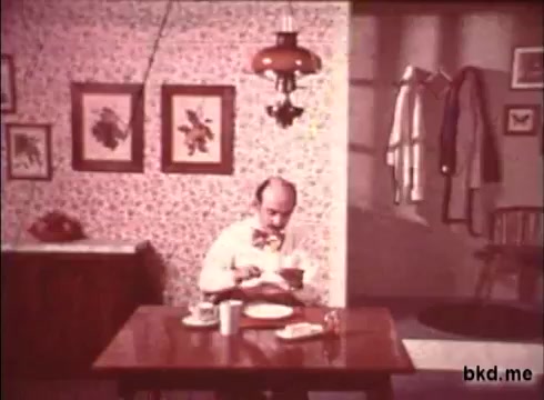 The still from 'Imperial Spread - 1970s' features a man sitting at a small wooden dining table in a modestly decorated room. He wears a bow tie and appears to be focused on a bowl in front of him. The walls are adorned with floral-patterned wallpaper and framed artworks, while a hanging lamp provides soft lighting. A couple of dishes and a drink are placed on the table, contributing to an everyday domestic scene.