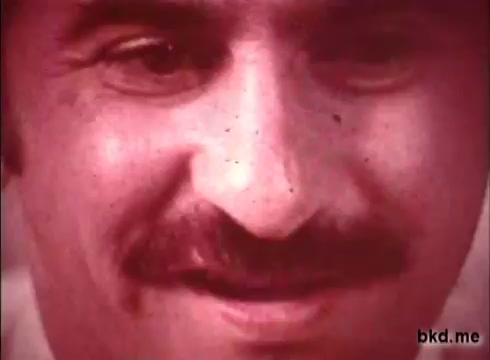 The still showcases a close-up of a man with a mustache, likely from the 1970s, characterized by the distinctive grain and color tone typical of 16mm film. The image conveys a warm yet muted atmosphere, reflecting the aesthetic of the time. The man's expression is contemplative, possibly smiling, engaging the viewer with a sense of nostalgia.