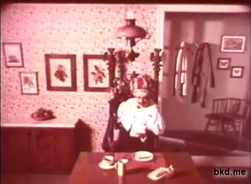 The still shows a vintage interior setting typical of the 1970s, featuring a dining table set with dishes and utensils. A figure, possibly a doll or a character, is at the table, dressed in period-appropriate clothing. The walls exhibit floral wallpaper and framed artworks, adding to the nostalgic ambiance. A lamp hangs above, casting a warm light over the scene. A coat rack and a chair are visible in the background, contributing to the homey atmosphere.