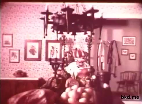 The still from 'Imperial Spread - 1970s' features a domestic interior scene characterized by vintage decor. The room has patterned wallpaper adorned with framed artwork. A prominent feature is a hanging light fixture with multiple arms. In the foreground, there's a variety of fruits on a table, and a chair is visible in the background. The overall color palette reflects the aesthetics of the era, with soft, muted tones.
