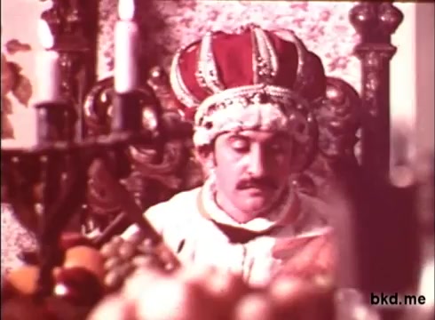 The still depicts a scene from 'Imperial Spread,' featuring a regal figure adorned in traditional royal attire and a lavish crown. He sits on an ornate throne, surrounded by sumptuous fruits, suggesting a setting of opulence and power. The warm color palette typical of 1970s films enhances the scene's richness. The atmosphere conveys a sense of authority and majesty.