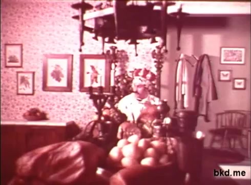 The still from 'Imperial Spread - 1970s' depicts a cozy interior scene. A man, dressed in period attire, is seated at a table laden with fruits, primarily oranges. The background features a patterned wallpaper, framed pictures, and a coat hanging nearby, contributing to a nostalgic atmosphere typical of the era. The overall color palette is warm and inviting, reflecting the style of 1970s cinema.