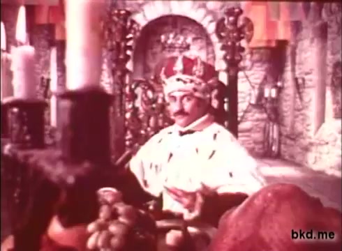The still depicts a regal figure seated on a throne, adorned in elaborate royal attire, including a crown. The setting features ornate architectural elements and rich textures, suggesting a historical or fantastical context. The color palette appears vibrant, typical of 1970s film aesthetics. The composition conveys a sense of authority and grandeur.