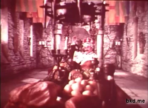 The still from 'Imperial Spread - 1970s' features a richly decorated dining hall with stone walls and banners. In the foreground, a lavish display of food, including meats and fruits, is prominently arranged. The central focus appears to be a figure seated on a throne-like chair, likely representing a regal character. The overall atmosphere suggests opulence and grandeur, typical of historical or fantasy-themed settings. The colors are vibrant, reflecting the distinct aesthetic of 16mm film from that era.