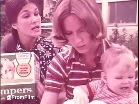 The still from the 1970s Pamper's commercial features two women and a baby. One woman, dressed in a patterned blouse, appears to be speaking animatedly, holding a box of Pampers. The second woman, wearing a striped shirt, looks concerned as she holds a visibly upset baby. The setting seems casual, possibly outdoors, reflecting the family-centric themes common in advertisements of that era. The vintage quality of the 16mm print adds a nostalgic feel to the image.