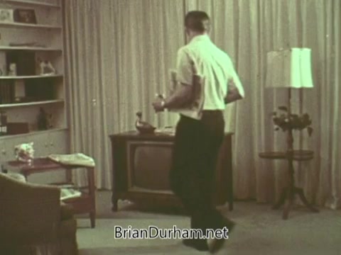 The still depicts a man in a light shirt carrying books while walking in a living room. The decor includes a vintage television, a table lamp, and a mid-century modern chair. The overall atmosphere is typical of a 1960s home setting, with curtains providing a soft backdrop. The scene suggests a focus on domestic life, possibly tied to the PSA's themes promoting community and church involvement.
