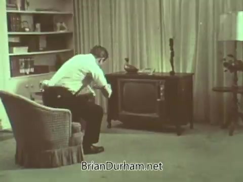 The still depicts a man sitting in a living room, facing a television set. He appears to be engaged with the screen, possibly watching a public service announcement. The decor suggests a typical 1960s household, with a cozy atmosphere highlighted by the furnishings and lighting. The overall color and tone reflect the era's aesthetic, contributing to the nostalgic feel of the scene.