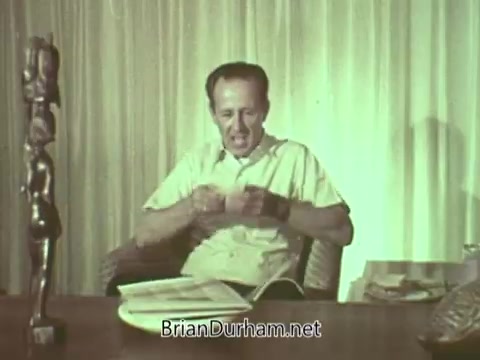 The still features a man seated at a desk, engaged in conversation or presentation. He appears to be in a casual setting, surrounded by decor items, including a statue. The background is a light curtain, suggesting a relaxed atmosphere. The focus may be on discussing topics related to the Episcopal Church, reflecting the themes of the PSA from the 1960s.