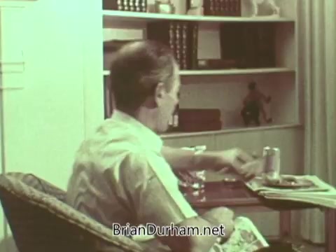 The still features an older man sitting in a wicker chair, eating and reading at a small table. The setting appears homely, with shelves in the background filled with books and decorative items. The overall tone suggests a casual, personal moment, likely highlighting themes of community or reflection in the context of the PSA.