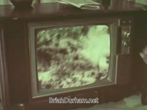 The still features a vintage television set displaying an indistinct, blurry image that appears to be natural scenery, possibly with trees or foliage. The TV is positioned on a wooden stand with some items placed on it, suggesting a home viewing environment typical of the 1960s. The overall aesthetic is characterized by the era’s design and color palette.