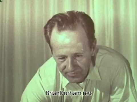 The still shows a man in a light-colored shirt, seemingly engaged in a thoughtful or serious conversation. He appears to be looking down, possibly at a document or object in front of him. The background has a simple, draped fabric that suggests a studio setting. The overall tone reflects a mid-20th century aesthetic typical of public service announcements from that era.