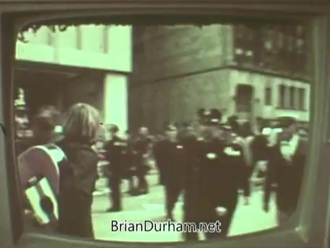 The 16mm still from the 1960s Episcopal Church PSA shows a scene on a city street, with a crowd of people, including law enforcement officers, visible in the foreground. The image captures a moment of social interaction or event, likely highlighting a community or public issue relevant to the church's outreach during that era. The frame displays a vintage television quality, emphasizing its historical context.
