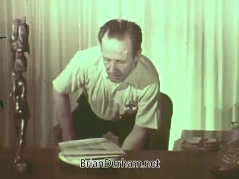 The still from the 1960s Episcopal Church PSA features a man seated at a desk, appearing contemplative as he leans over a book or document. The setting includes a sculpture and some books, suggesting a reflective or serious atmosphere. The color palette has a muted quality indicative of the era's film style.