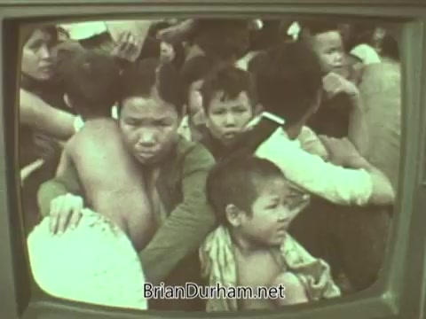 The still depicts a crowded scene, possibly reflecting a moment of distress. It shows a group of people, including children, with expressions of fear and concern. The image is captured within a vintage television frame, suggesting it is a still from a public service announcement. The overall tone conveys a sense of urgency and highlights the plight of those depicted.