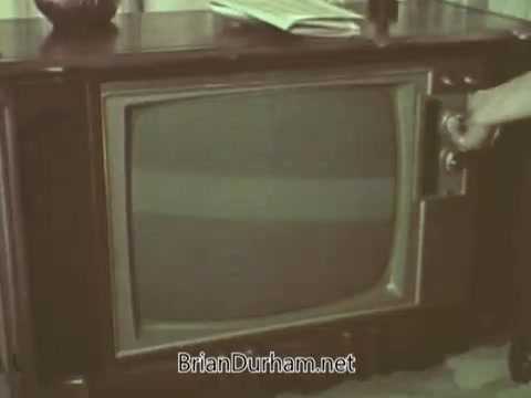 The still shows a person reaching to adjust an older television set, typical of the 1960s. The TV is housed in a wooden cabinet and displays horizontal lines on the screen, suggesting it may be turned off or experiencing reception issues. In the background, there's a glimpse of a table with a decorative item and some papers, adding context to a domestic setting. The overall tone is faded, indicative of the era.