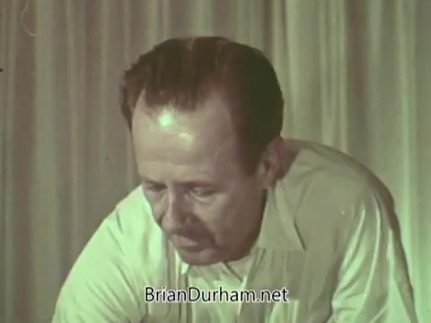 The still shows a man focused intently on a task, with a neutral expression. He appears to be dressed in a light-colored shirt, and the background consists of a plain curtain. The overall tone of the image suggests a serious or reflective moment, characteristic of public service announcements from the 1960s.