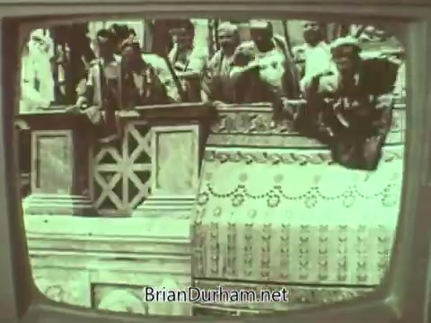 The still from the 'Episcopal Church PSA - 1960s' depicts a scene featuring a group of people, likely members of the church, gathered around a balcony or raised platform. They appear to be engaged in a ceremonial or public event, with decorative elements in the background. The image is likely captured in a vintage 16mm format, giving it an aged aesthetic typical of that era.