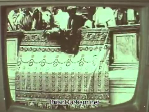 The still from the 'Episcopal Church PSA - 1960s' features a vintage television display with a monochrome image. It depicts a group of people gathered, likely in a church setting, with a prominent patterned cloth or banner in the foreground. The atmosphere suggests a ceremonial or communal event, reflective of church activities during that era. The image conveys a sense of tradition and community within the Episcopal Church context.