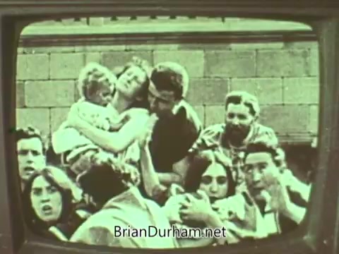 The still features a group of people gathered closely together, some appearing distressed. A woman holds a child, while others engage with each other, showing a range of emotions. The scene is presented in a vintage television frame, reminiscent of the 1960s. The overall atmosphere suggests themes of community and concern, typical of a public service announcement.