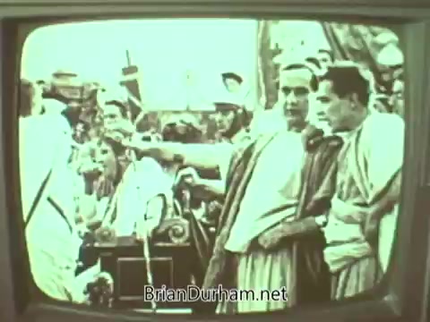 The still from the 'Episcopal Church PSA - 1960's' shows a group of men dressed in period costumes, likely from a historical reenactment or biblical scene. They appear to be engaged in conversation, with one man gesturing, suggesting a moment of decision or discussion. The scene captures a vintage aesthetic, typical of mid-20th century film, and may convey themes relevant to the Episcopal Church's message during that era. The monochrome quality adds to the historical context of the piece.