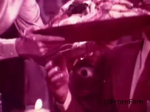 The still from the 'Coffee-Mate Commercial - Space Needle - 1970's' features a close-up shot of a dining scene. A hand is reaching out to present a tray of food, while another hand, presumably belonging to a man in a suit, is shown reaching for the tray. The color scheme is a vintage pinkish hue, characteristic of 1970s film aesthetics. The background hints at a lively atmosphere, possibly in a restaurant setting. The scene captures a moment of hospitality and indulgence, fitting for a coffee creamer commercial.