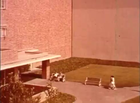 The still captures a quiet outdoor scene, likely from an educational film focused on the human cell and cytology. The setting includes a landscaped area with a bench, where two individuals are seated, while a child walks by. The backdrop features a large brick building, suggesting a medical or educational environment. The overall tone is calm and conducive to a learning atmosphere.