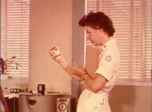 The still features a nurse in a crisp white uniform, examining a small container in her hands. She stands in a laboratory setting, with a countertop appliance visible behind her, likely a centrifuge or storage unit. Natural light filters through blinds, illuminating the scene, which conveys a focused and educational atmosphere typical of instructional films on cellular biology.