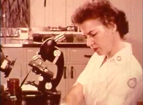 The still features a female cytologist working in a laboratory setting. She is focused on her tasks, likely preparing or analyzing specimens under a microscope. The background includes laboratory equipment and organizational elements typical of a scientific workspace. This scene is likely intended to illustrate the role of a cytologist in studying human cells, emphasizing the hands-on work involved in cytology.