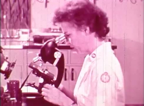 The still features a woman in a lab coat observing a microscope in a laboratory setting. The image is characterized by a vintage aesthetic typical of 16mm film, with a slight pink hue. Laboratory equipment and a cluttered workspace can be seen in the background, emphasizing the educational context of cytology.