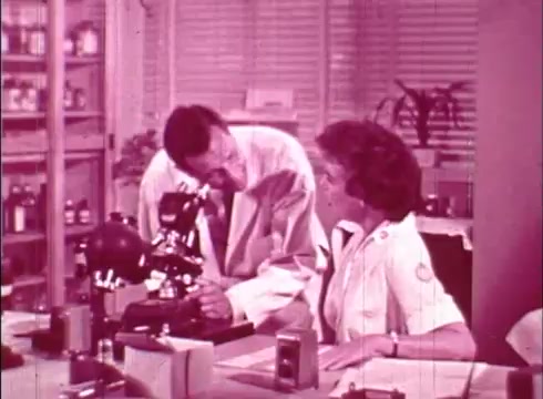 The still depicts a scene from an educational film focused on cell biology. It shows two individuals, a man and a woman, in a laboratory setting. The man, dressed in a lab coat, leans toward a microscope, appearing to discuss or demonstrate something to the woman, who is seated and wearing a nurse's uniform. The environment is filled with lab equipment and supplies, reflecting a scientific atmosphere. The overall color tone of the image is a soft pink, typical of 16mm prints from that era.