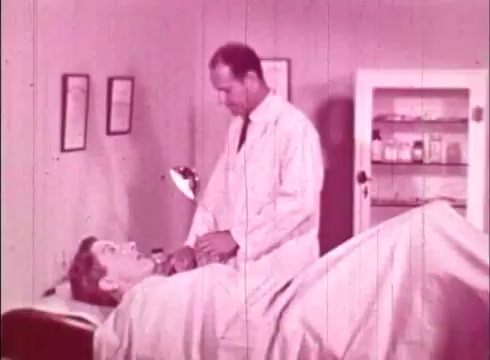 The still from the educational film 'The Human Cell and the Cytologist' depicts a medical examination setting. A man, presumably a patient, is lying on an examination table covered with a sheet, looking up at a healthcare professional. The professional, dressed in a white lab coat, appears to be preparing to conduct a procedure or examination. The background features medical charts on the wall and a cabinet filled with medical supplies, emphasizing the clinical environment. The overall color tone is a faded pink, characteristic of 16mm prints.