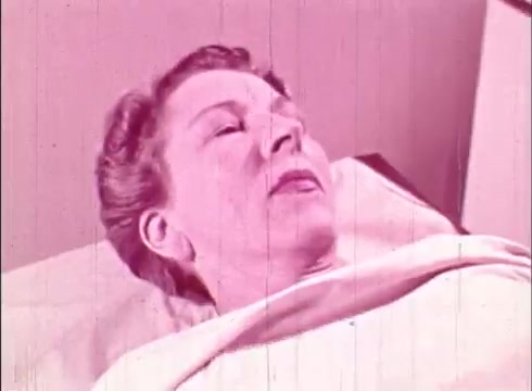 The still from 'The Human Cell and the Cytologist' depicts a woman lying in a sterile medical environment, possibly in a hospital or laboratory setting. The image has a pink tint, characteristic of some 16mm film prints. Her expression suggests a state of contemplation or focus, likely related to a scientific examination or discussion about cytology. The overall aesthetic conveys an educational context, emphasizing the human connection to cellular study.