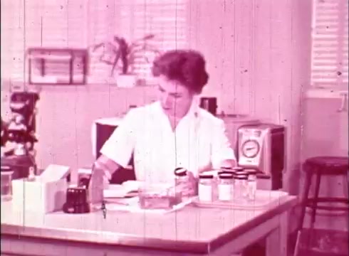 The still depicts a woman in a laboratory setting, engaged in her work with various glass containers and equipment on a table. She is focused on a task, possibly analyzing samples. The environment is typical of a cytology lab, with scientific instruments and a plant visible in the background. The color tone is pinkish, characteristic of older 16mm film prints, which often exhibit color fading. This image likely serves an educational purpose, showcasing the work of a cytologist.