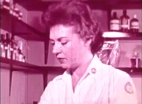 The still from 'The Human Cell and the Cytologist' depicts a woman in a lab setting, wearing a white lab coat. She appears focused on her work, with shelves filled with various bottles and lab supplies in the background. The image has a vintage aesthetic typical of 16mm film, with a soft pink hue and visible film grain, emphasizing its educational context.