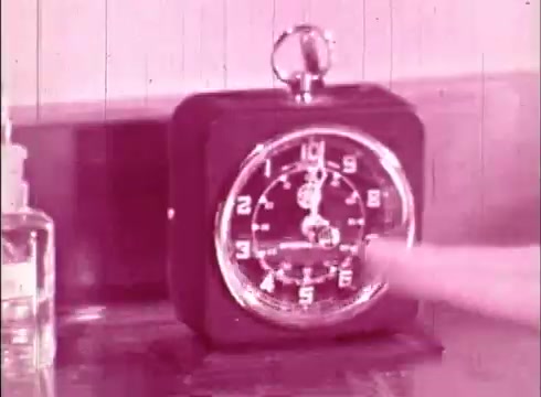 The still features a vintage alarm clock on a table, with a finger pointing towards the clock's face. The image has a pinkish hue typical of older 16mm prints. The setting suggests a moment of emphasis or timing that may relate to a lesson or demonstration in the educational film 'The Human Cell and the Cytologist.'